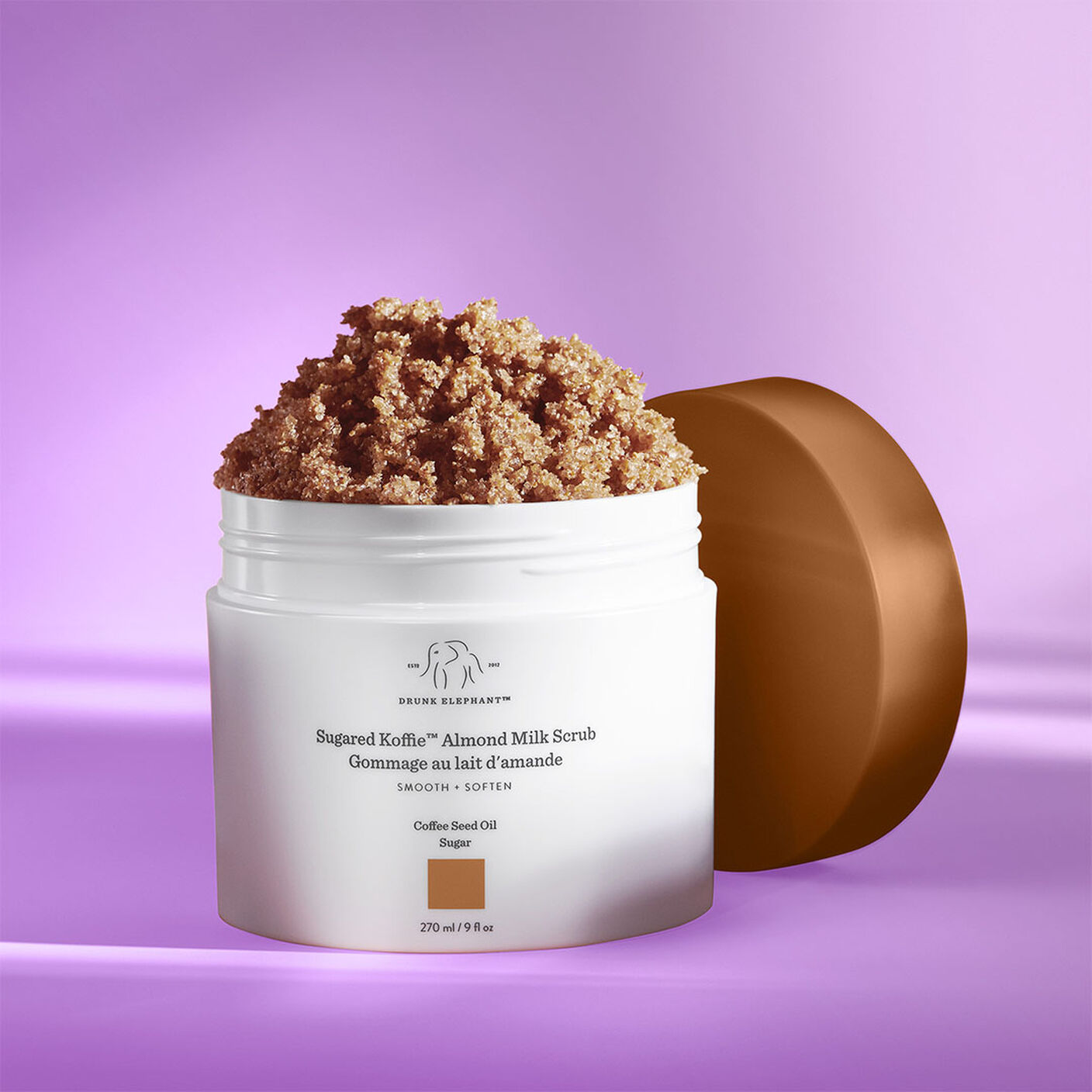 Gommage au lait d’amande sucré Koffie avec le bouchon enlevé sur une table mauve.