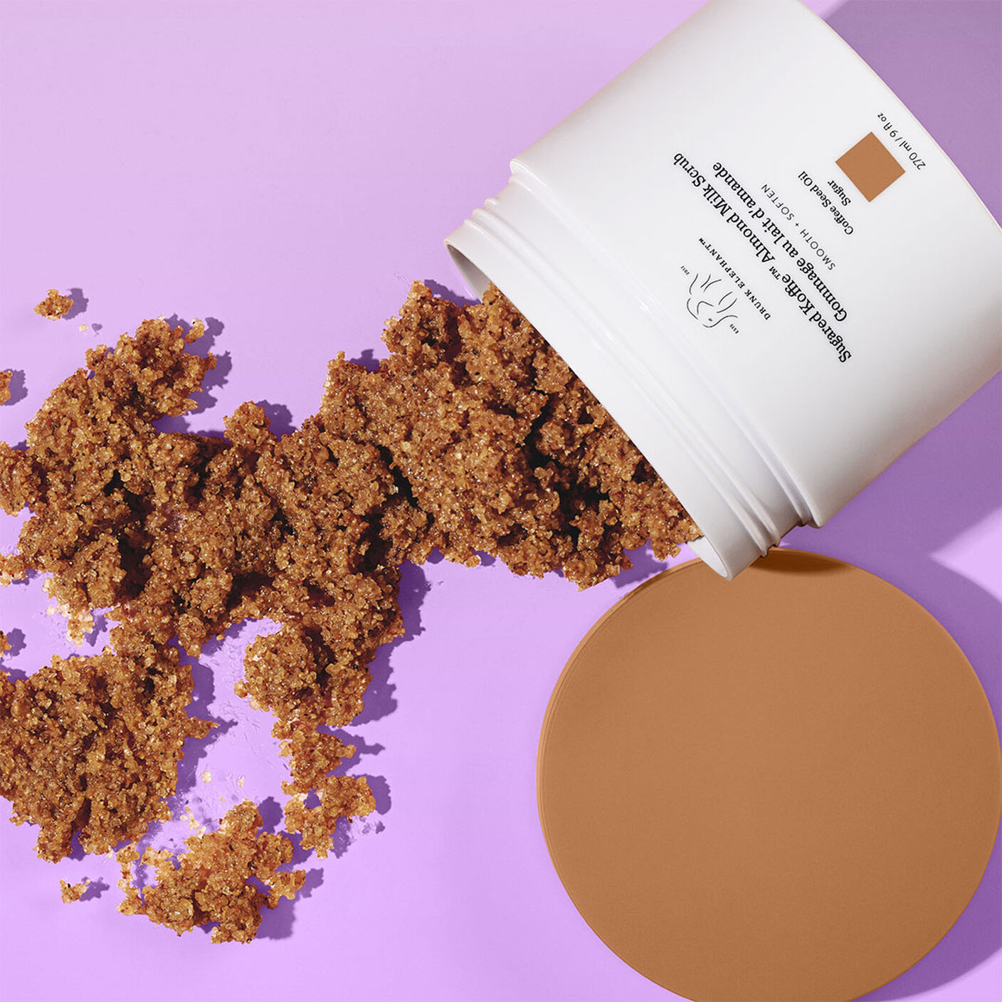 Gommage au lait d’amande sucré Koffie versé sur une table mauve avec le bouchon sur le côté.